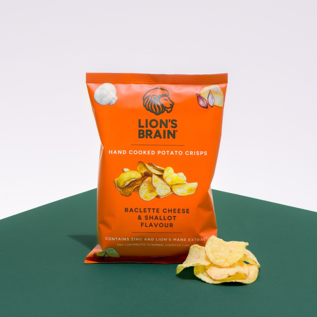 A bag of Lion's Brain Hand Cooked Potato Crisps in Raclette Cheese & Shallot Flavour, displayed on a dark green surface against a white background with a few crisps scattered in front.