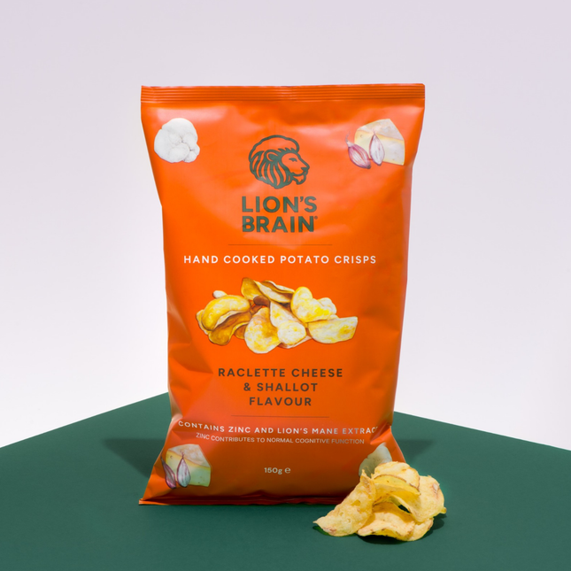 An orange bag of Lion's Brain hand cooked potato crisps, labelled 'Raclette Cheese & Shallot Flavour', standing on a dark green surface against a light grey background with a few crisps scattered in front.