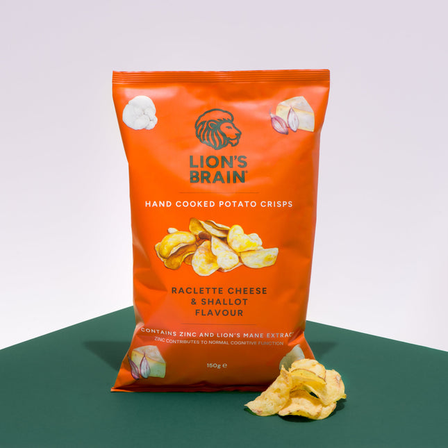 A bag of Lion's Brain hand cooked potato crisps in 'Raclette Cheese & Shallot' flavor, standing on a green surface against a white background with a few crisps placed in front of the bag.